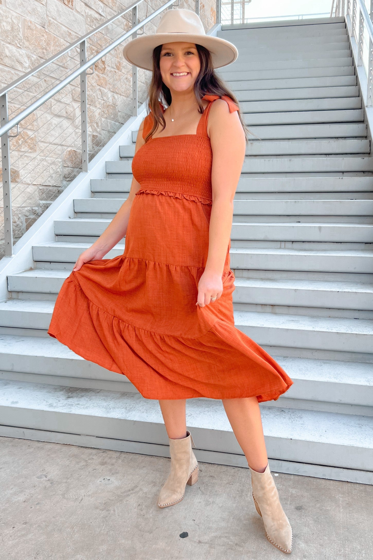 woman wearing an orange maternity dress with a smocked bodice and tiered skirt, holding her baby bump, on outdoor stairs