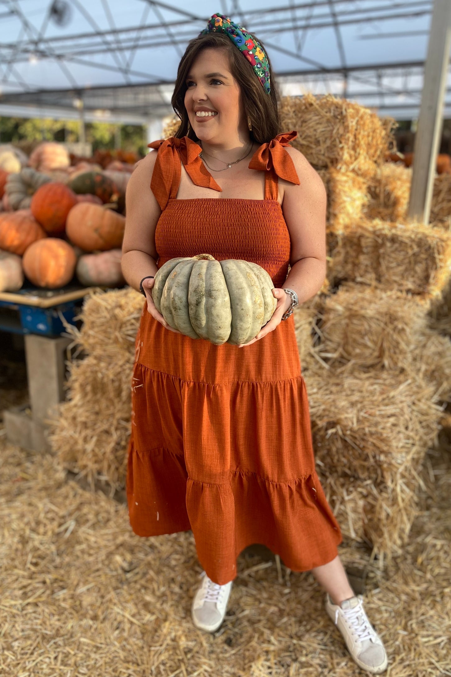 woman wearing an orange maternity dress with tie straps, holding a pumpkin in a pumpkin patch