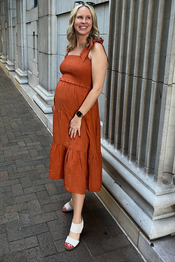 woman wearing an orange maternity sundress with ruffled tiers and a baby bump, standing against a stone wall