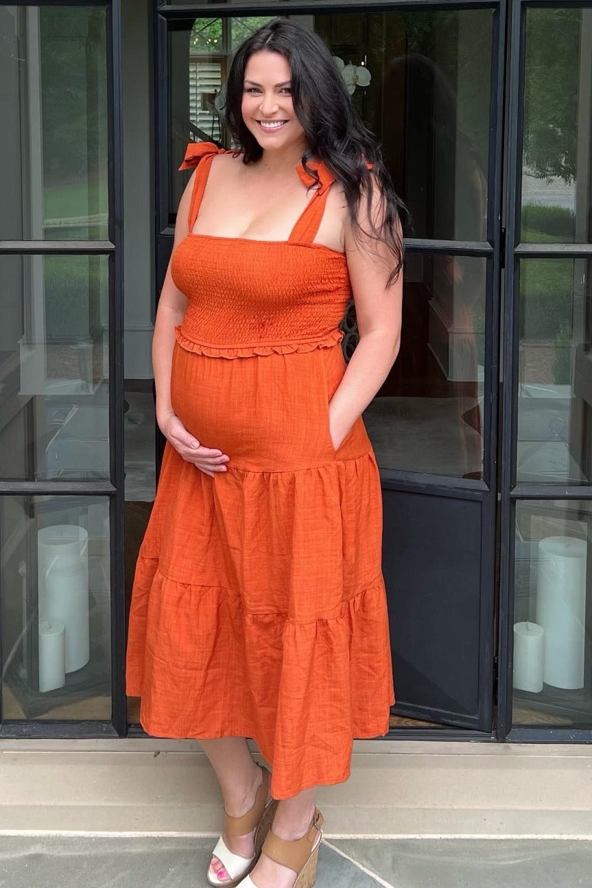 woman wearing an orange maternity dress with a smocked top and tiered skirt, holding her baby bump