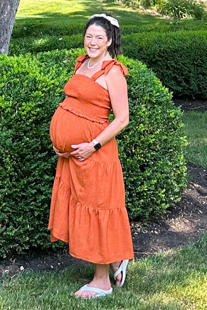 woman wearing an orange maternity dress with tiered detailing and holding her baby bump in a garden