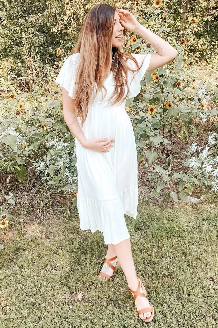 woman wearing a white maternity dress with flutter sleeves and a tiered hem, holding her baby bump in a garden