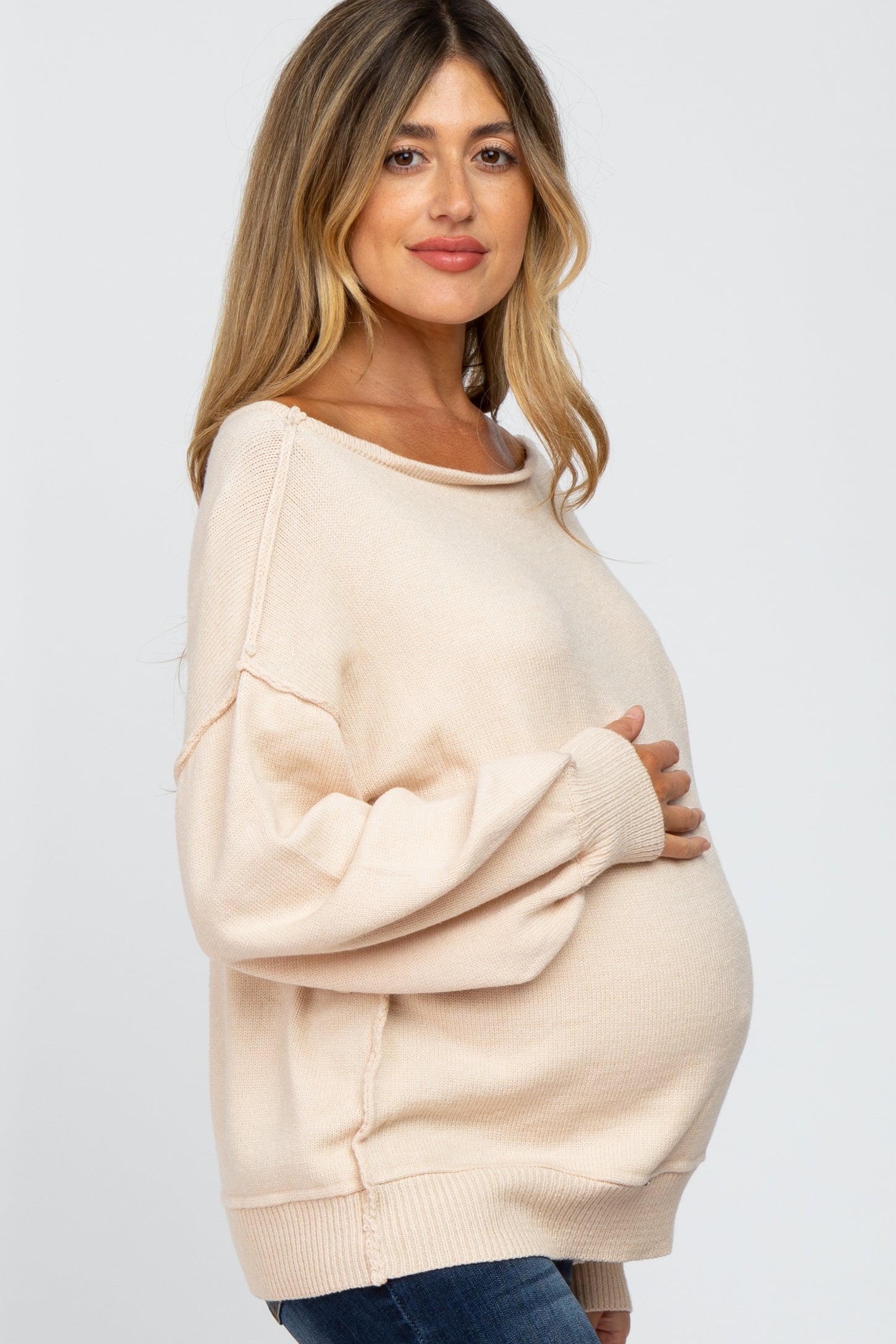 Pregnant woman in a cream oversized maternity sweater holding her baby bump against a white background