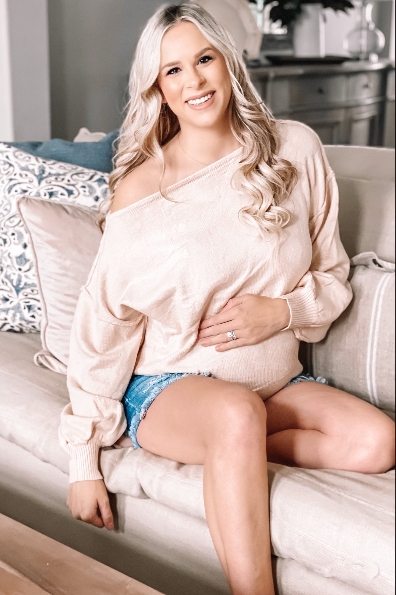 woman wearing a light beige off-shoulder maternity sweater and denim shorts, sitting on a couch and showing her baby bump