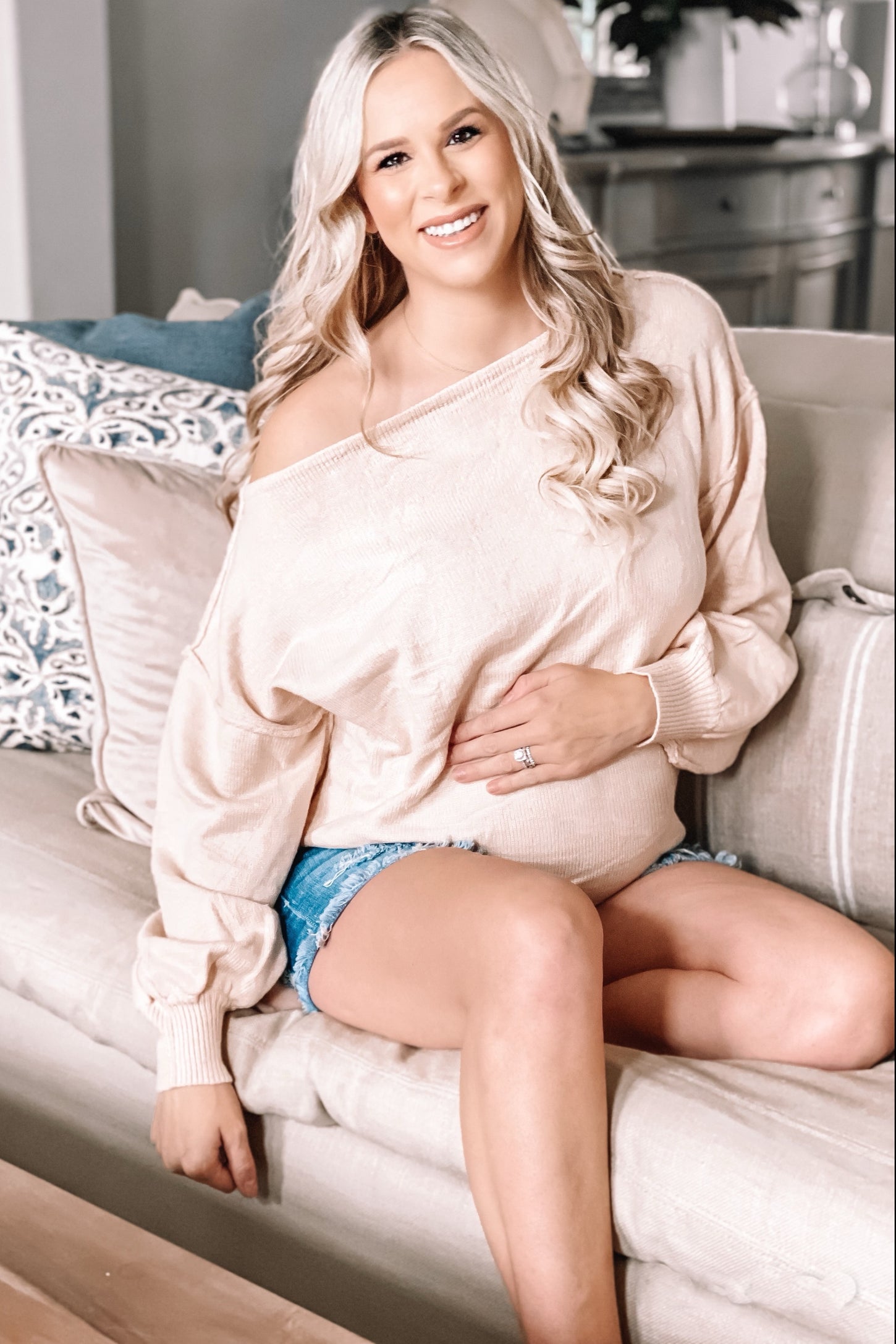 woman wearing a light beige off-shoulder maternity sweater and denim shorts, sitting on a couch and showing her baby bump