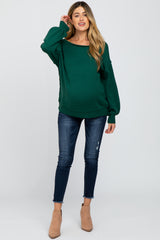woman wearing a green oversized maternity sweater and blue skinny jeans, showing her baby bump