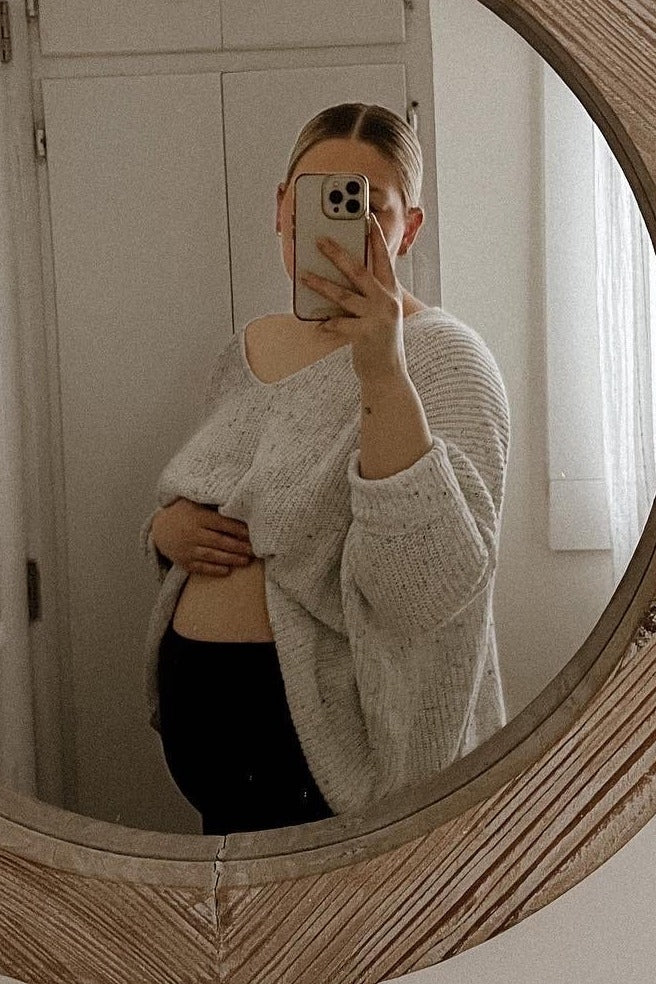 woman wearing a light gray oversized maternity sweater and holding her baby bump in front of a mirror