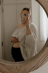woman wearing a light gray oversized maternity sweater and holding her baby bump in front of a mirror