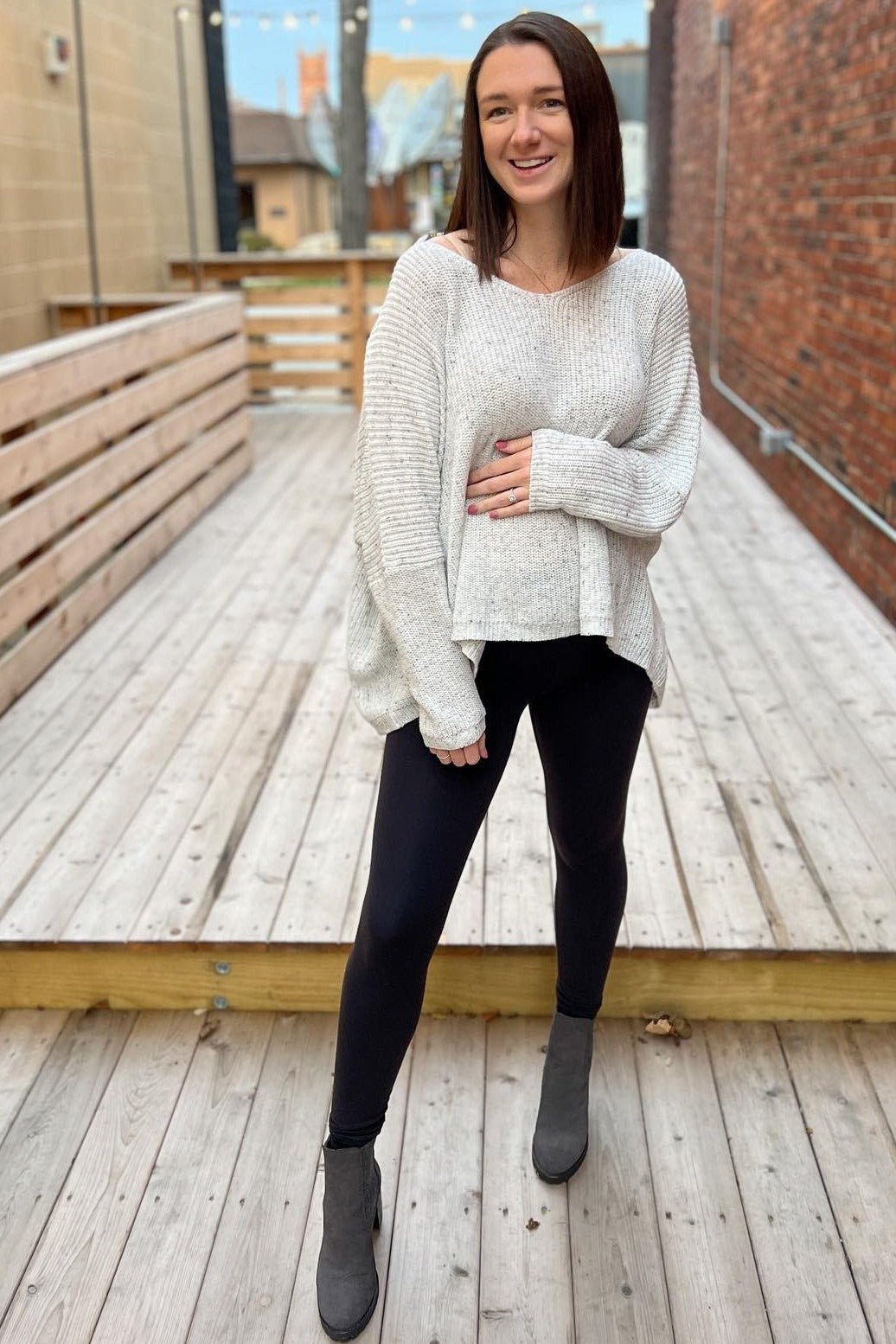 woman wearing a light gray oversized maternity sweater and black leggings, gently holding her baby bump
