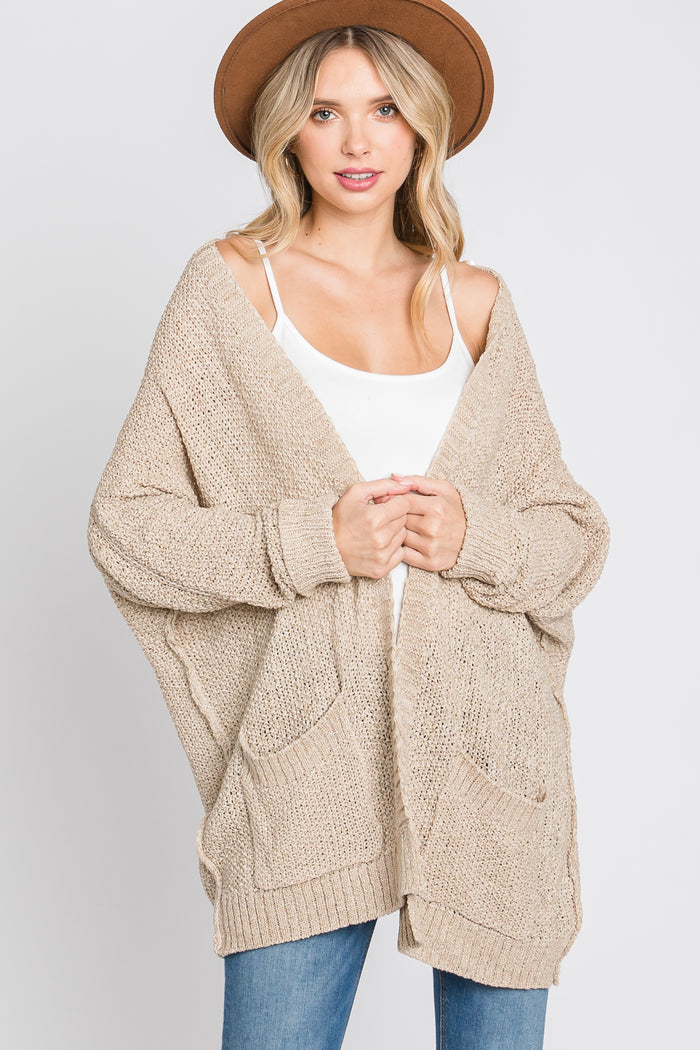 woman wearing a beige oversized knit cardigan with pockets and a white tank top, styled with a brown hat