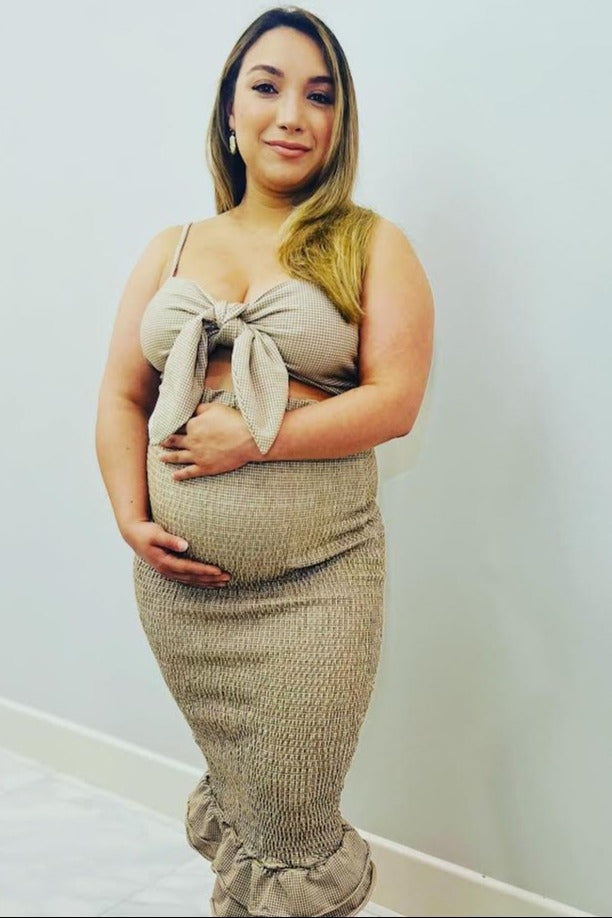 woman wearing a beige smocked maternity dress with a tie front and ruffle hem, holding her baby bump