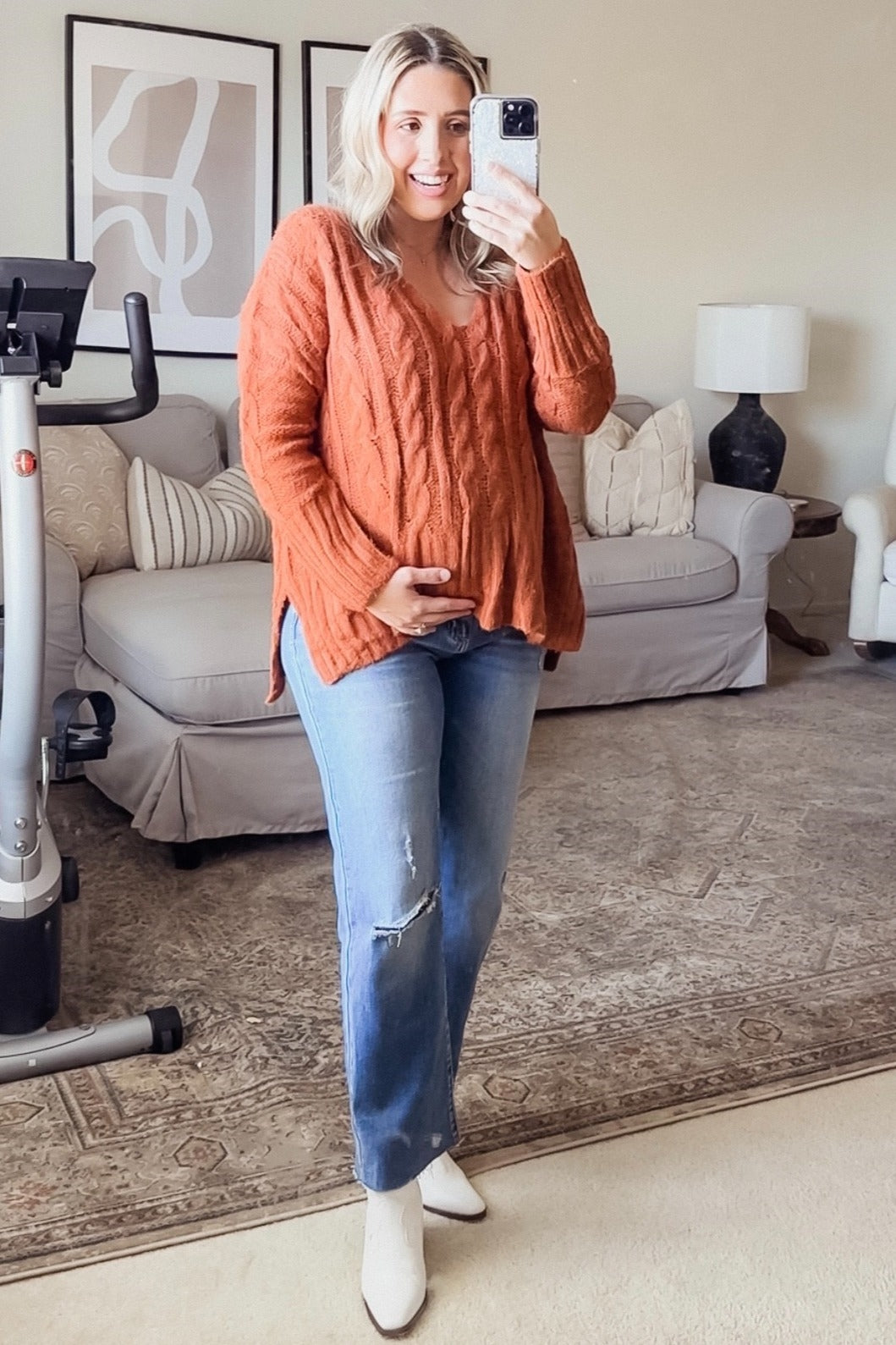 woman wearing an orange cable knit maternity sweater and holding her baby bump, paired with blue distressed jeans