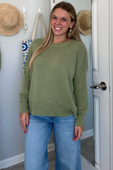 woman wearing a green oversized sweater and light blue wide-leg jeans, smiling in front of a mirror