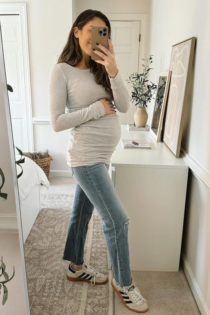 woman wearing a light gray long sleeve maternity top and jeans, holding her baby bump in front of a mirror