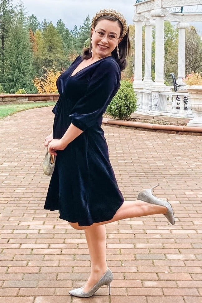 woman wearing a navy velvet wrap dress with three-quarter sleeves and silver high heels, smiling outdoors