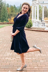 woman wearing a navy velvet wrap dress with three-quarter sleeves and silver high heels, smiling outdoors