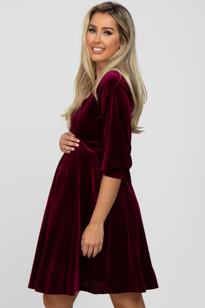 woman wearing a burgundy velvet maternity dress, holding her baby bump with a smile
