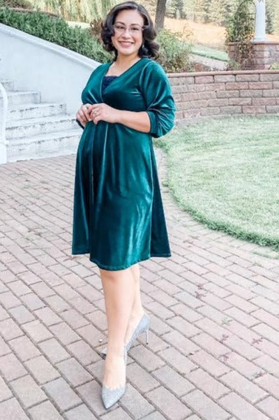 woman wearing a dark green velvet wrap dress and showing her baby bump while standing outdoors on a brick path