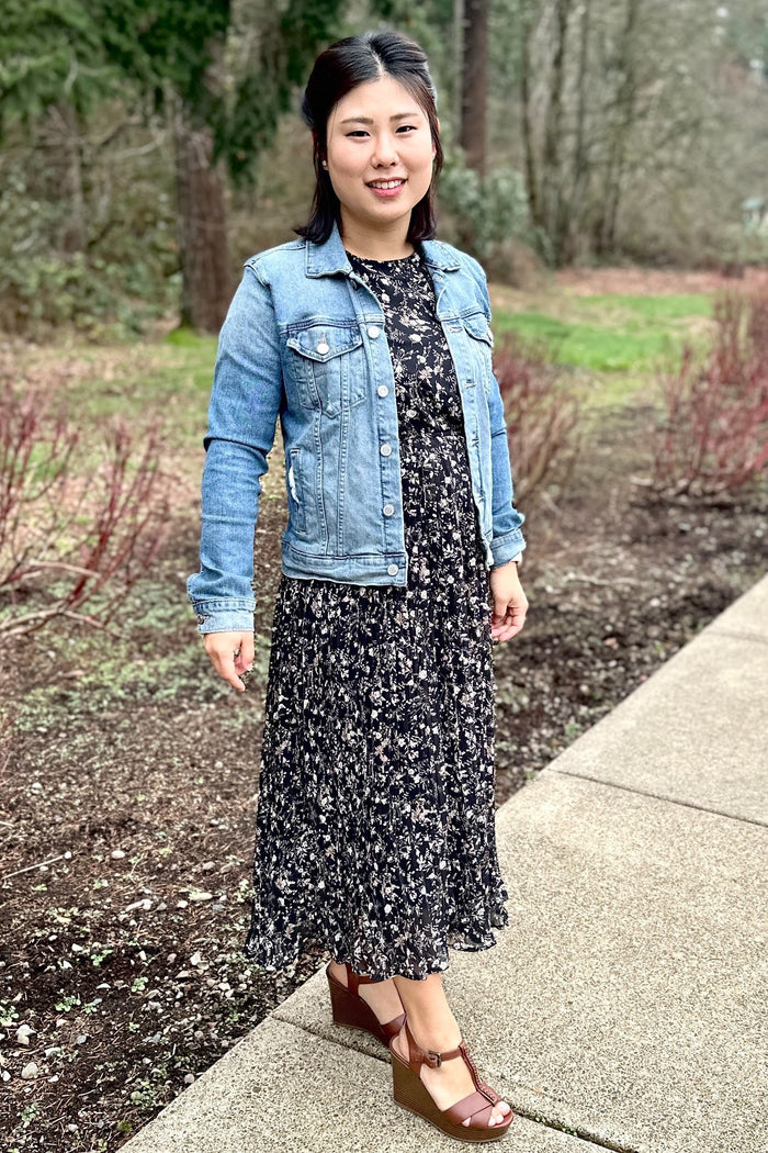woman wearing a black floral maxi dress, denim jacket, and brown wedge sandals, standing outdoors