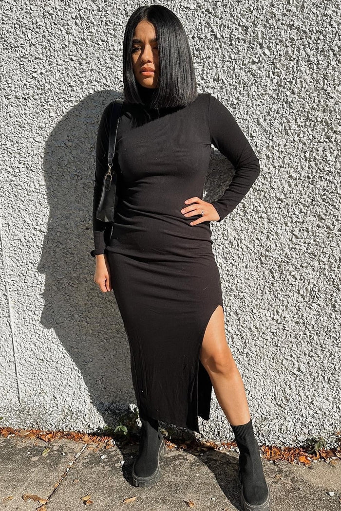 woman wearing a black long sleeve dress with a side slit and black ankle boots, standing against a textured wall