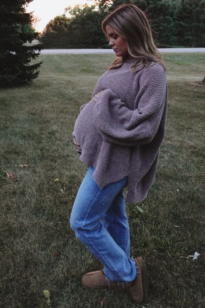 woman wearing a taupe oversized maternity sweater and blue jeans, cradling her baby bump in an outdoor setting