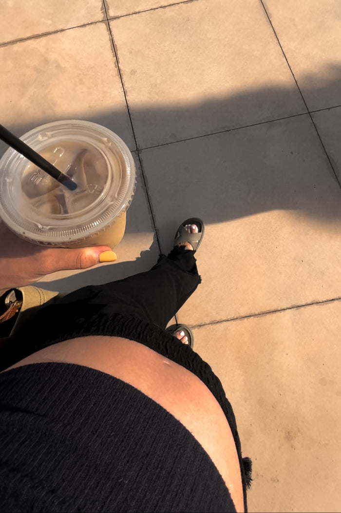 woman wearing a black maternity top and black ripped jeans, holding an iced coffee, with a visible baby bump