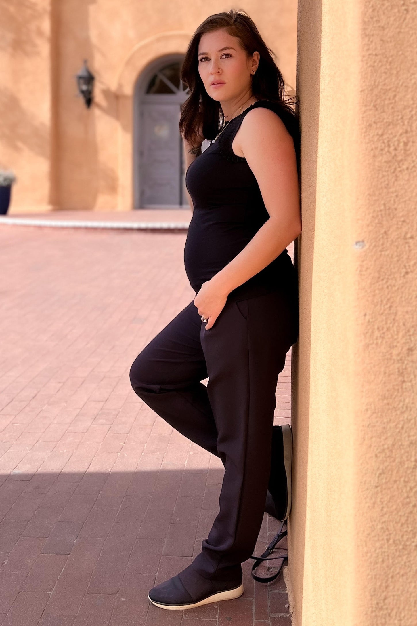 woman wearing a black maternity tank top and black maternity joggers, showing her baby bump while leaning against a wall