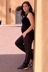 woman wearing a black maternity tank top and black maternity joggers, showing her baby bump while leaning against a wall