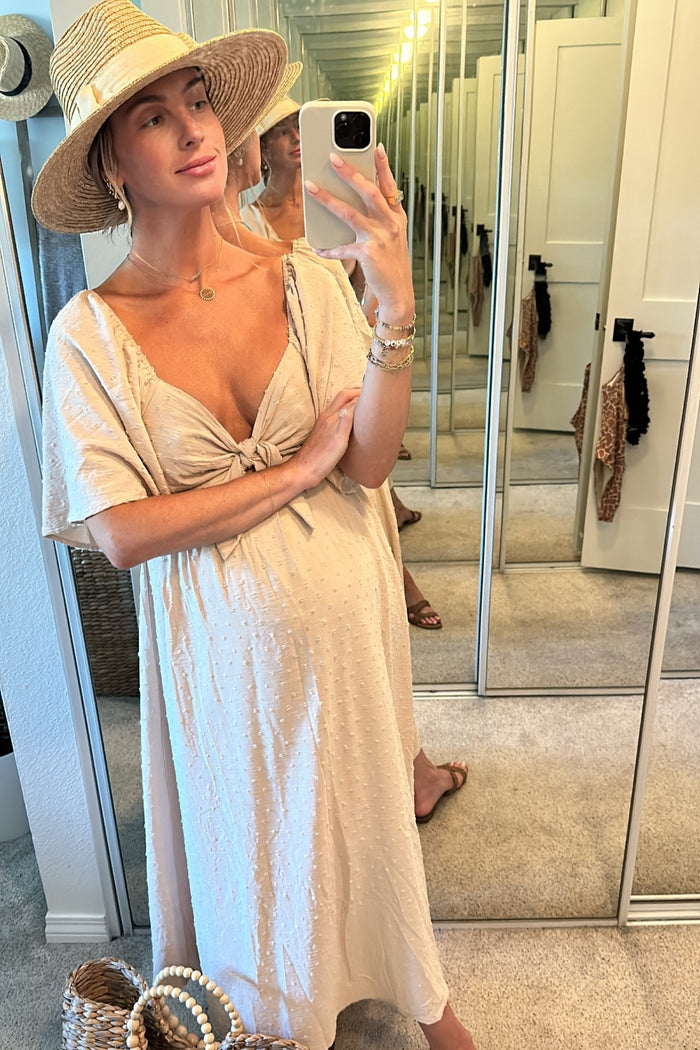 woman wearing a light beige maternity dress with a knotted front and polka dot texture, holding a straw hat and baby bump visible