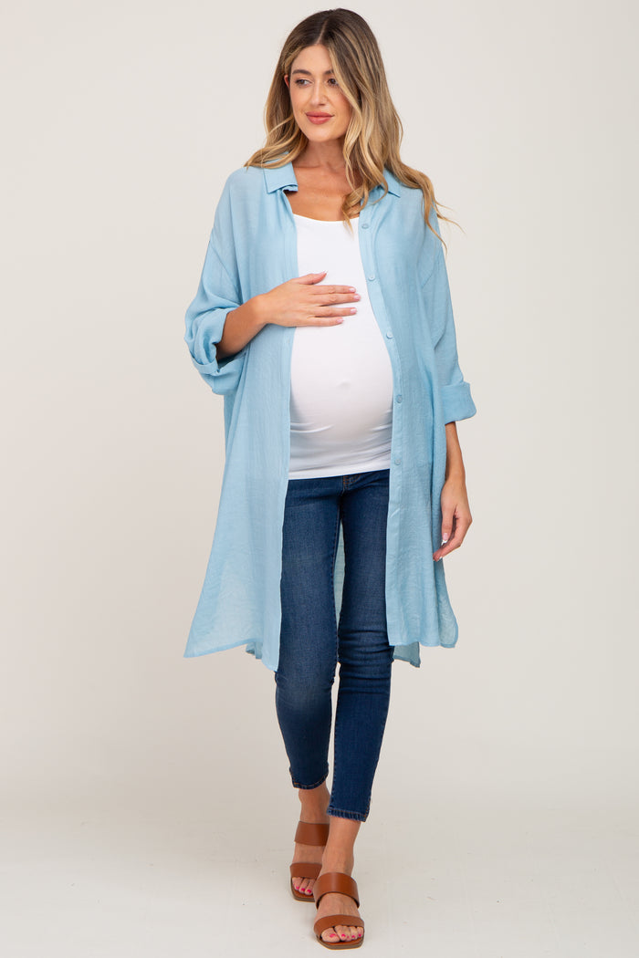 woman wearing a light blue maternity cover-up over a white top and showing her baby bump while standing in jeans