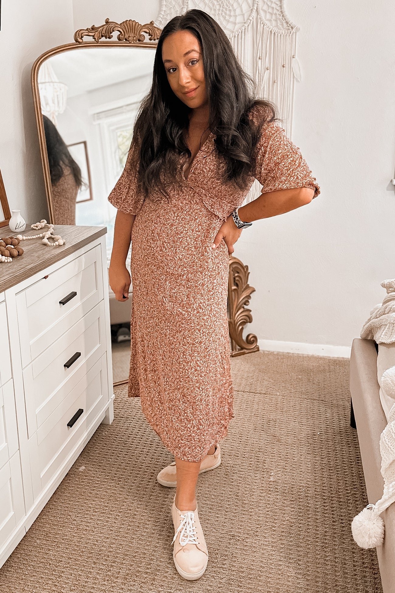 woman wearing a pink floral maternity dress and white sneakers, posing with one hand on her hip and a visible baby bump