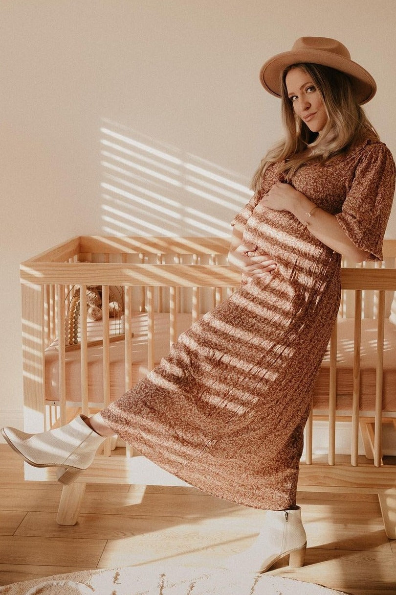 woman wearing a floral maternity dress and a hat, cradling her baby bump next to a wooden crib