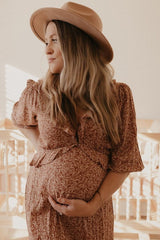 woman wearing a floral brown maternity dress and holding her baby bump, accessorized with a beige hat