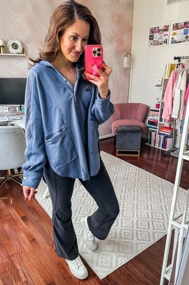 woman wearing a blue zip-up hoodie with pockets and black flared pants, standing in a bright room with a pink chair