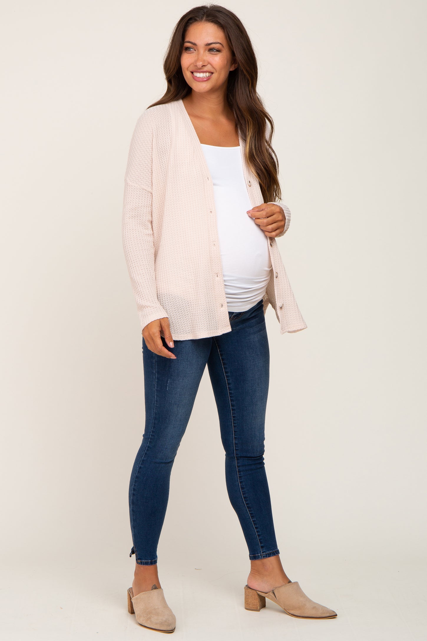 woman wearing a light pink maternity cardigan over a white top and blue maternity jeans, showing her baby bump