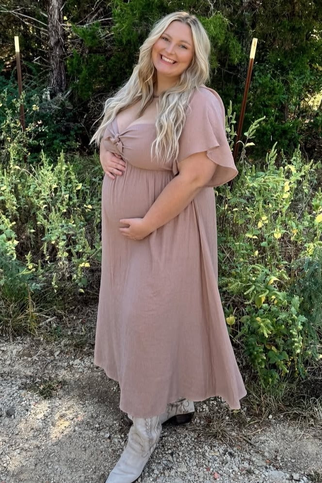 Smiling pregnant woman in a mauve maternity maxi dress and white cowboy boots posing outdoors