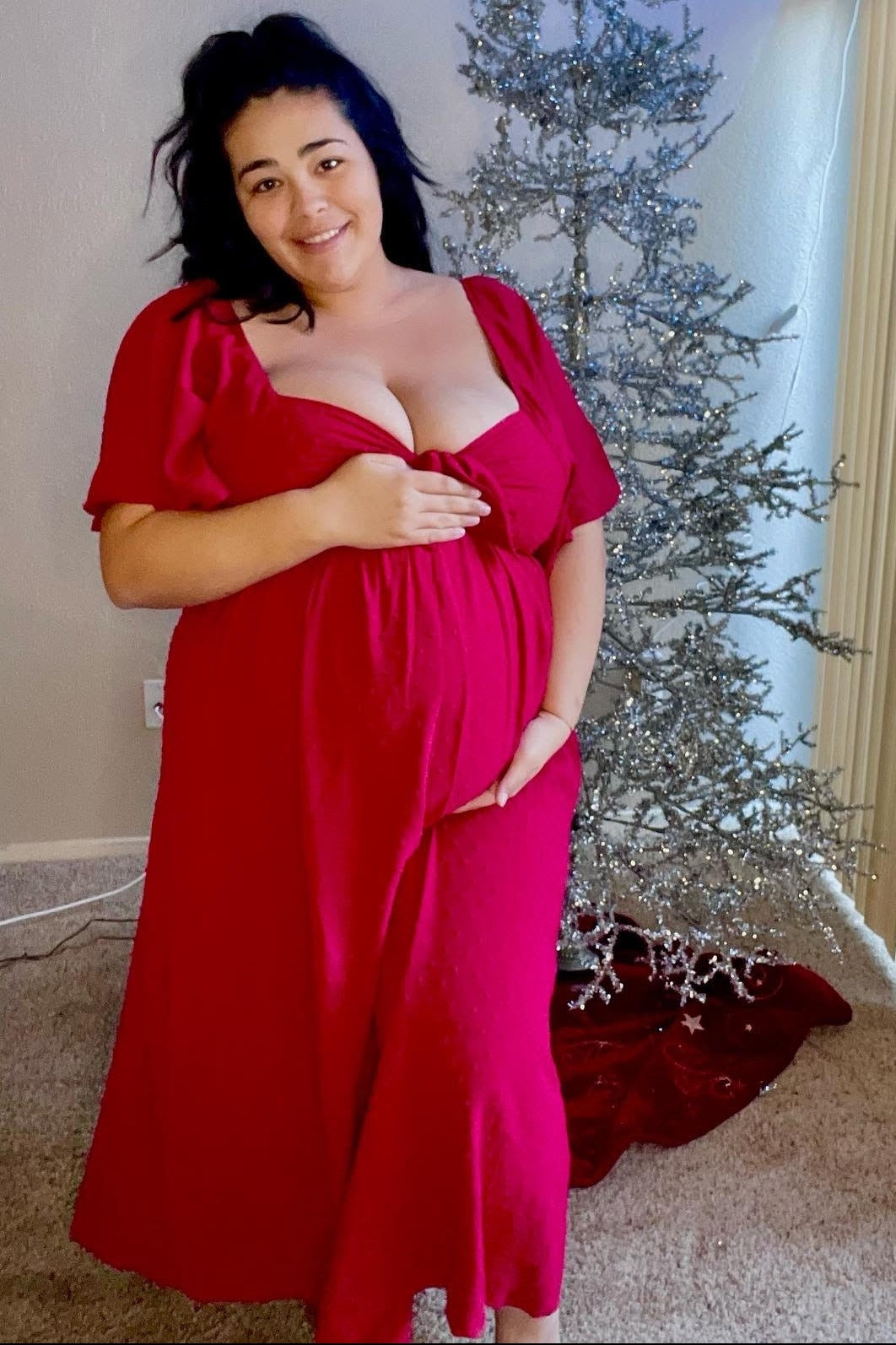 woman wearing a bright red maternity dress with short sleeves and a baby bump, standing next to a beautifully decorated Christmas tree