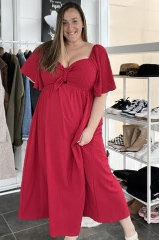 woman wearing a red maxi dress with a knot detail and flutter sleeves, standing in a stylish clothing boutique