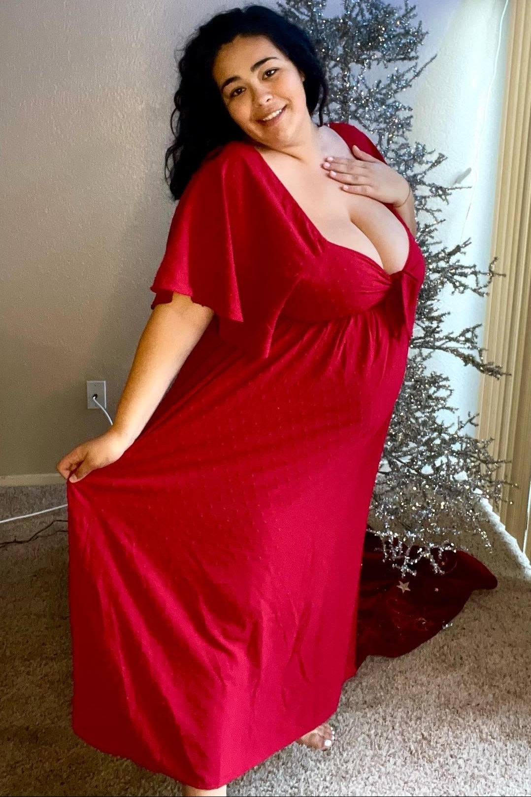 woman wearing a red maxi dress with flutter sleeves, posing with a smile in front of a decorative tree