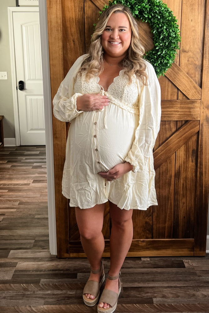 woman wearing a cream maternity dress with lace details and buttons, smiling and cradling her baby bump