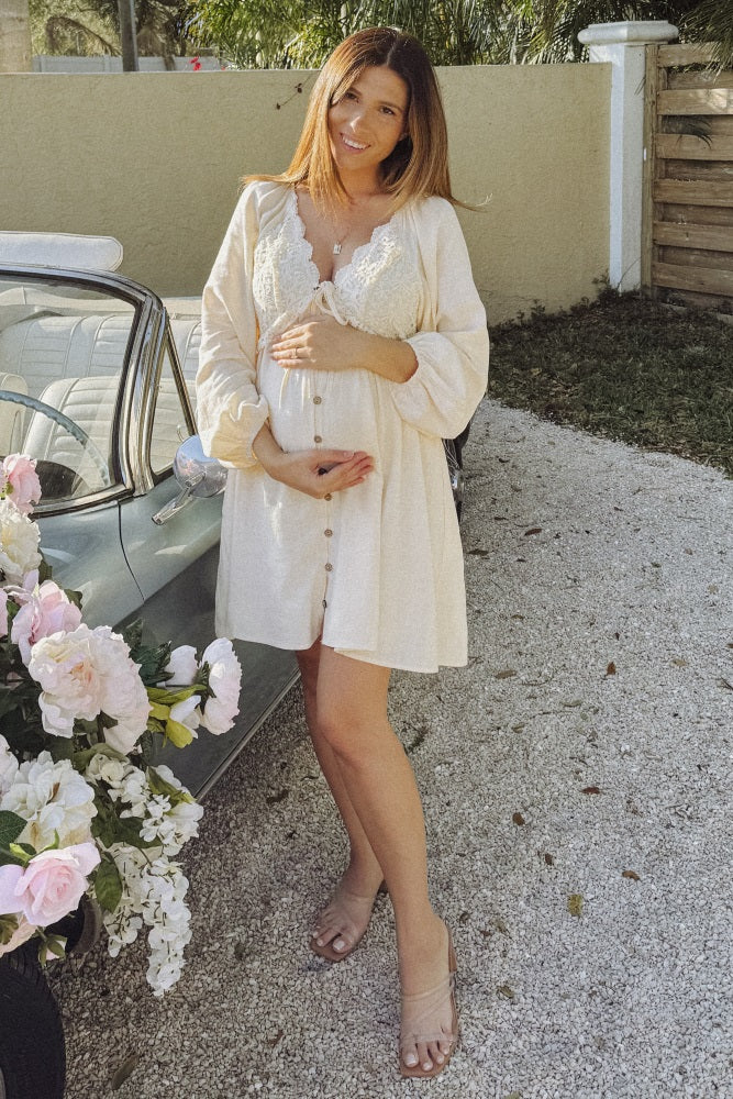 woman wearing a cream-colored maternity dress with lace details and a visible baby bump, standing next to a vintage car