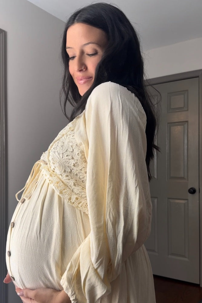 woman wearing a cream maternity dress with lace detailing, showing her baby bump while posing sideways