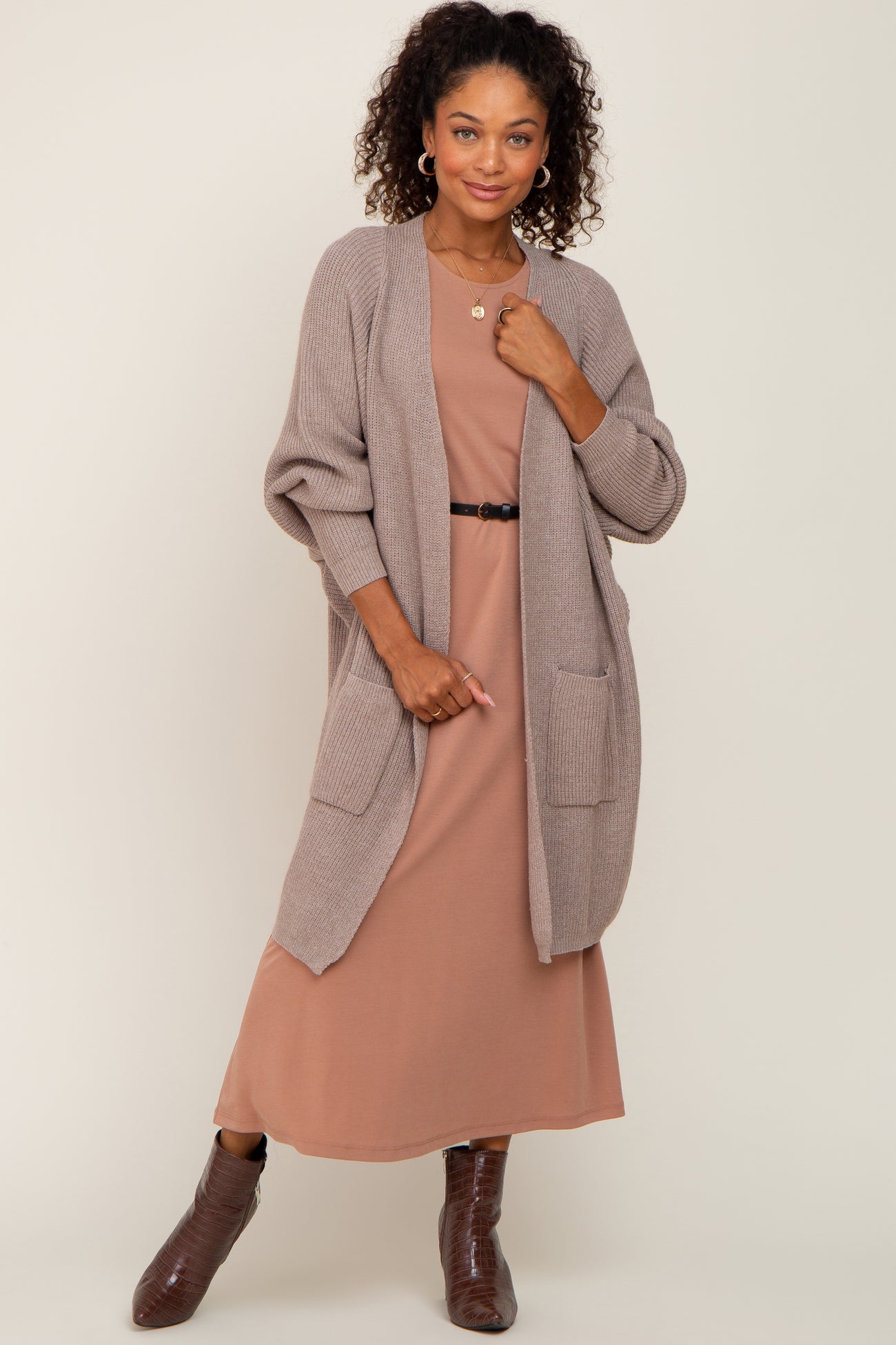 woman wearing a tan knit cardigan over a rose-colored midi dress with pockets and a black belt, posing stylishly