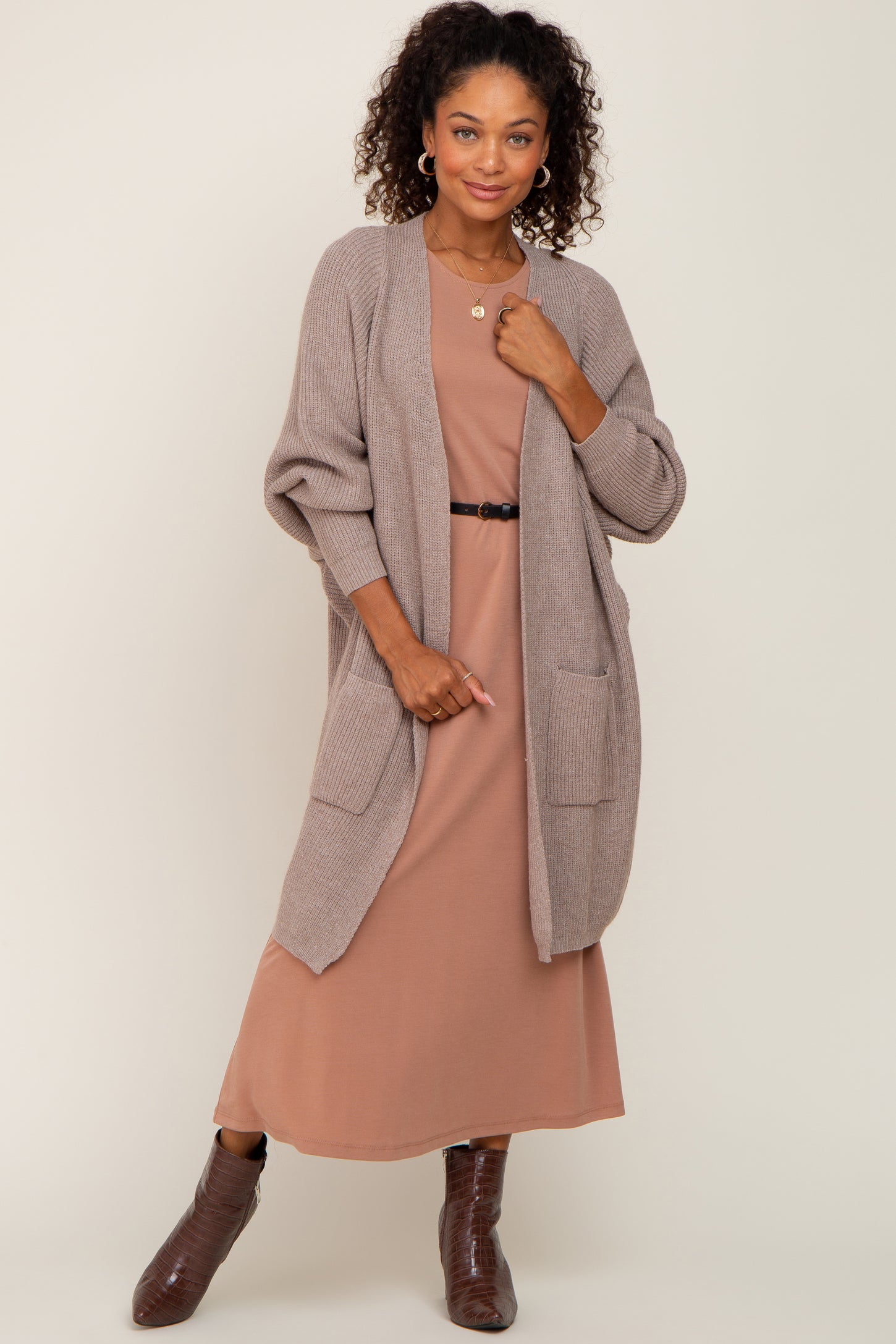 woman wearing a tan knit cardigan over a rose-colored midi dress with pockets and a black belt, posing stylishly