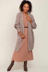 woman wearing a tan knit cardigan over a rose-colored midi dress with pockets and a black belt, posing stylishly