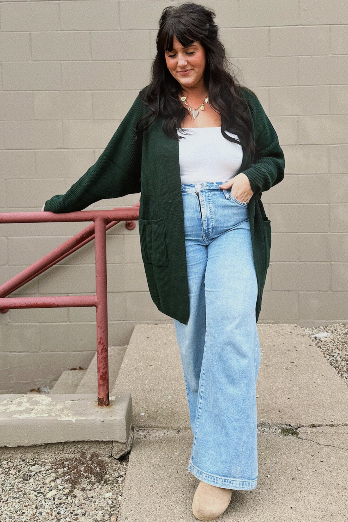 woman wearing a dark green long cardigan over a white tank top and light blue wide-leg jeans, standing by a railing