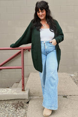 woman wearing a dark green long cardigan over a white tank top and light blue wide-leg jeans, standing by a railing