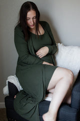 woman wearing a dark green ribbed maternity dress and cardigan, gently holding her baby bump while sitting on a couch