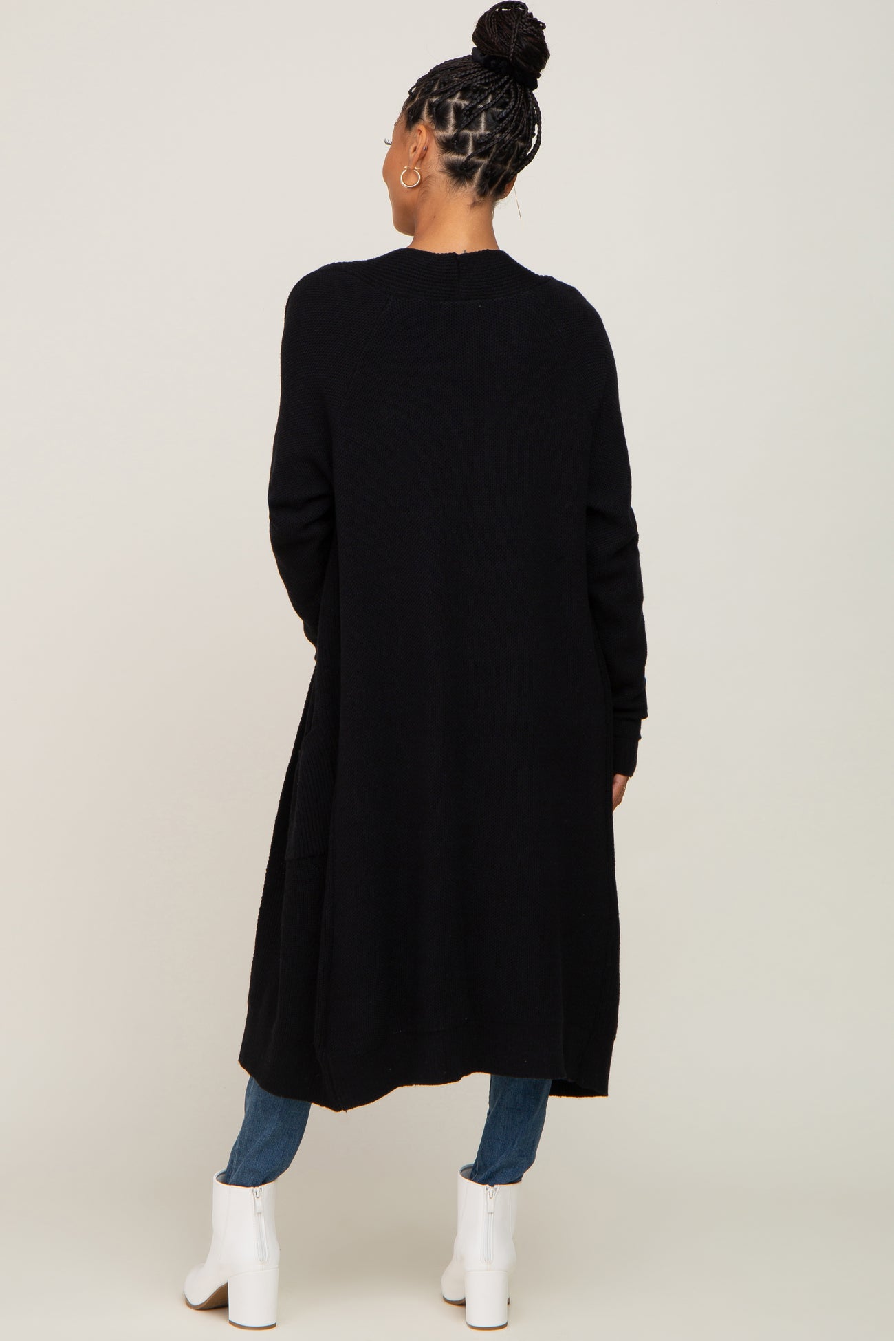 woman wearing a long black cardigan with an open back and pockets, styled with jeans and white ankle boots