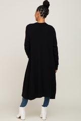 woman wearing a long black cardigan with an open back and pockets, styled with jeans and white ankle boots